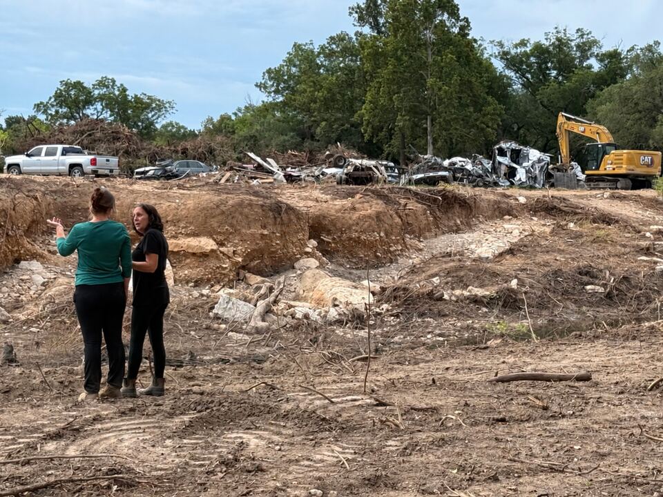 Two women standing on bare scoured ground where the creek cut a new channel, with an excavator and wrecked vehicles working along the far bank.