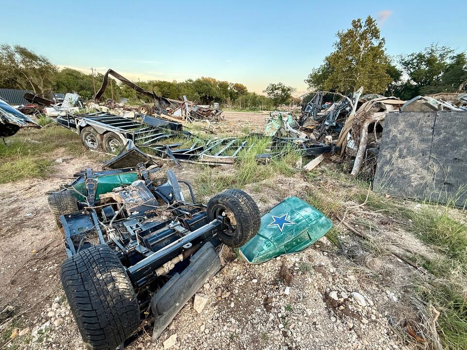 A golf cart lying upside down in the grass with a trailer chassis stripped to its frame beside it and a wall of twisted metal and timber piled behind, in low evening light.