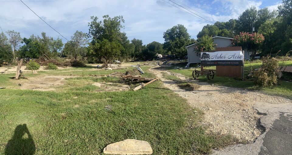 The same entrance after the flood. The fence and the
                  outbuildings are gone, the ground is scoured to bare dirt and
                  scattered with broken timber and debris, and the trees are
                  stripped. The painted Asher Acres sign is still standing at
                  the right of the drive.