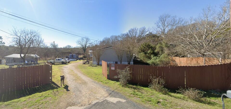 The front entrance of Asher Acres before the flood: a gravel
                  drive running between a stained wooden privacy fence and a
                  mown lawn, with the family dog standing in the drive, a
                  pickup truck and outbuildings beyond, and bare winter trees
                  along the treeline.