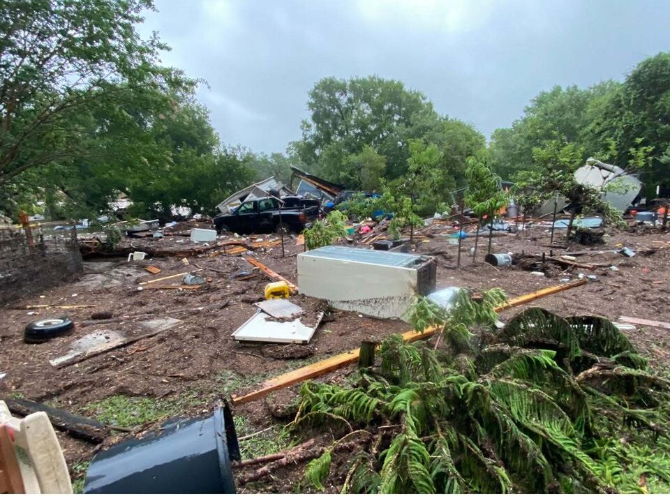 The orchard after the water went down: young fruit trees snapped and bent in churned mud, a refrigerator lying on its side, a crushed pickup and a collapsed outbuilding beyond, under a grey sky.