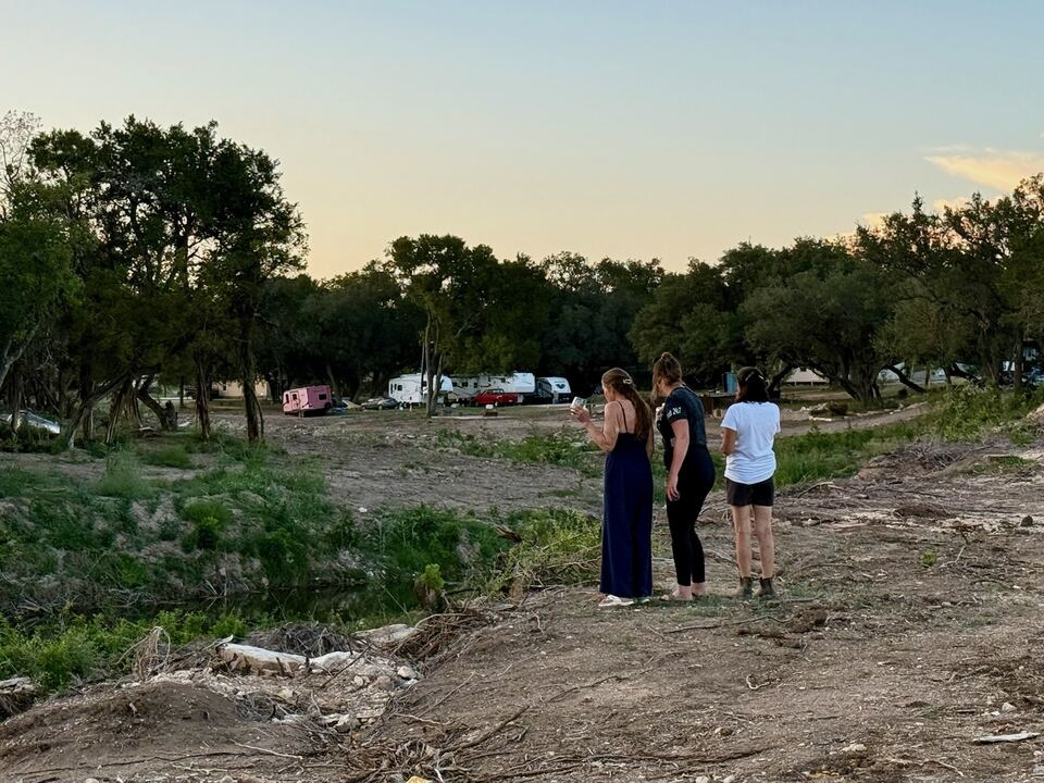 Brandy, Donna and Ashlee standing together at dusk with
                    their backs to the camera, looking out across bare scoured
                    ground toward a row of travel trailers drawn up under the
                    trees on the far side.