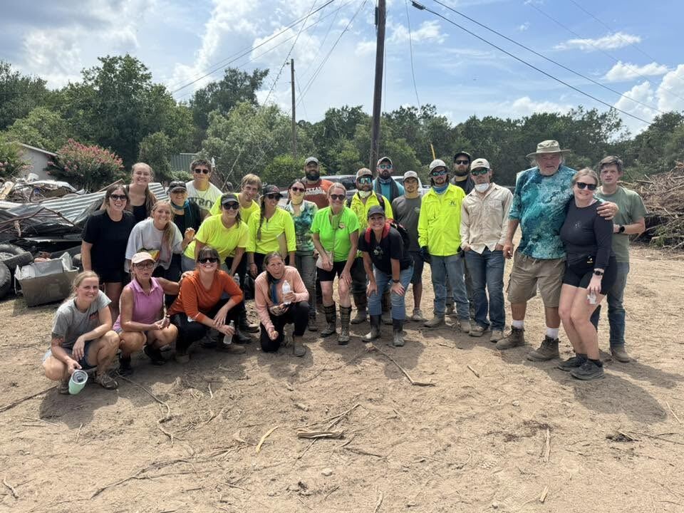 About twenty volunteers gathered together on the cleared
                  property, most in high-visibility vests and hard hats, with
                  piled flood debris and heavy equipment behind them.