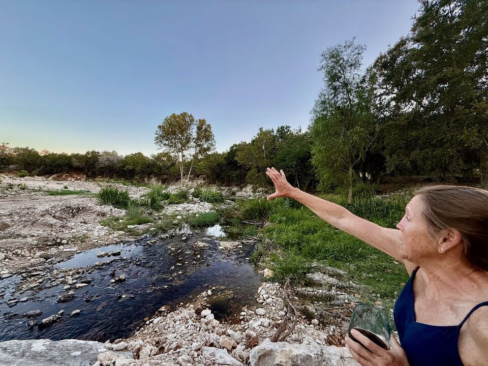Brandy Gerstner standing at the edge of Sandy Creek at
                  dusk, one arm raised to point out across the water and the
                  scoured limestone bed toward the trees on the far bank.