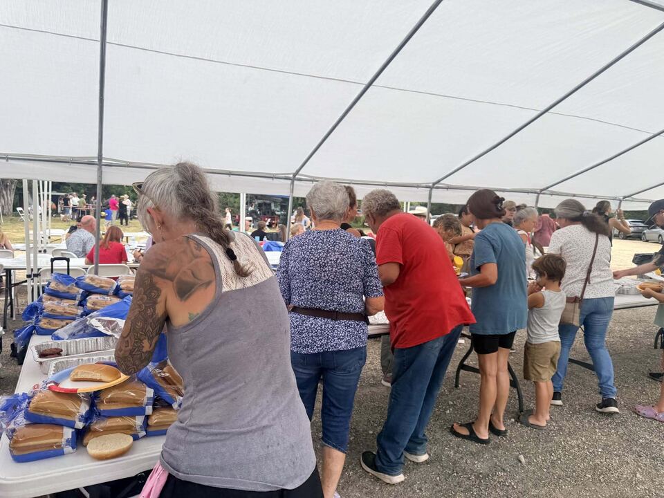 A community meal under a large open-sided event tent. Trestle
                  tables are lined with sliced bread and foil trays, neighbours
                  are serving and collecting food, and a small child waits in
                  the line.