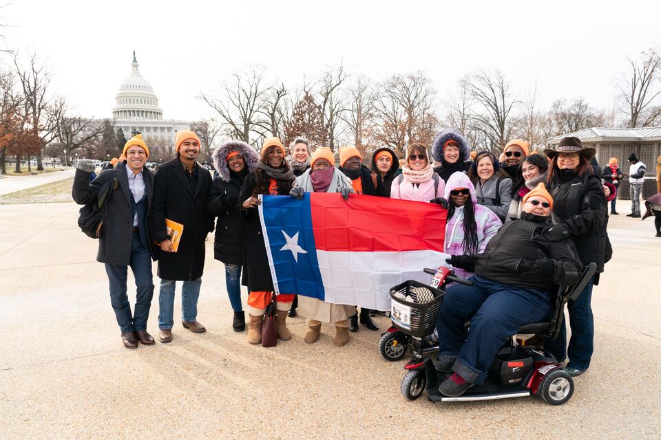 About twenty people from Sandy Creek and other Central Texas
                  communities standing together outside the United States
                  Capitol in winter coats and matching orange hats, holding a
                  Texas flag between them.