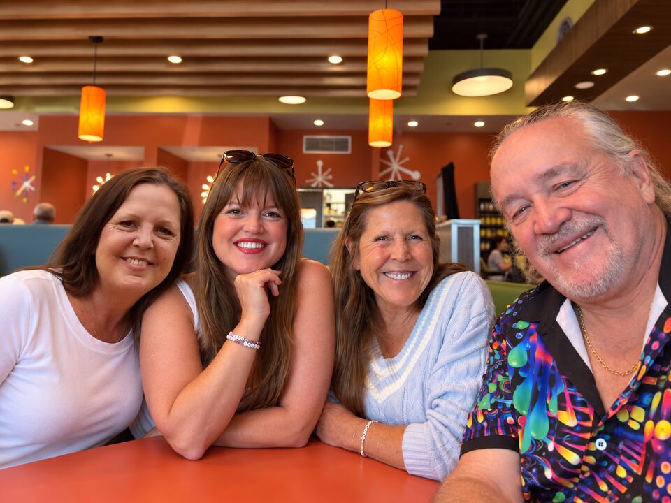 Four members of the family leaning in together around a
                  table in a brightly lit diner, all smiling at the camera.