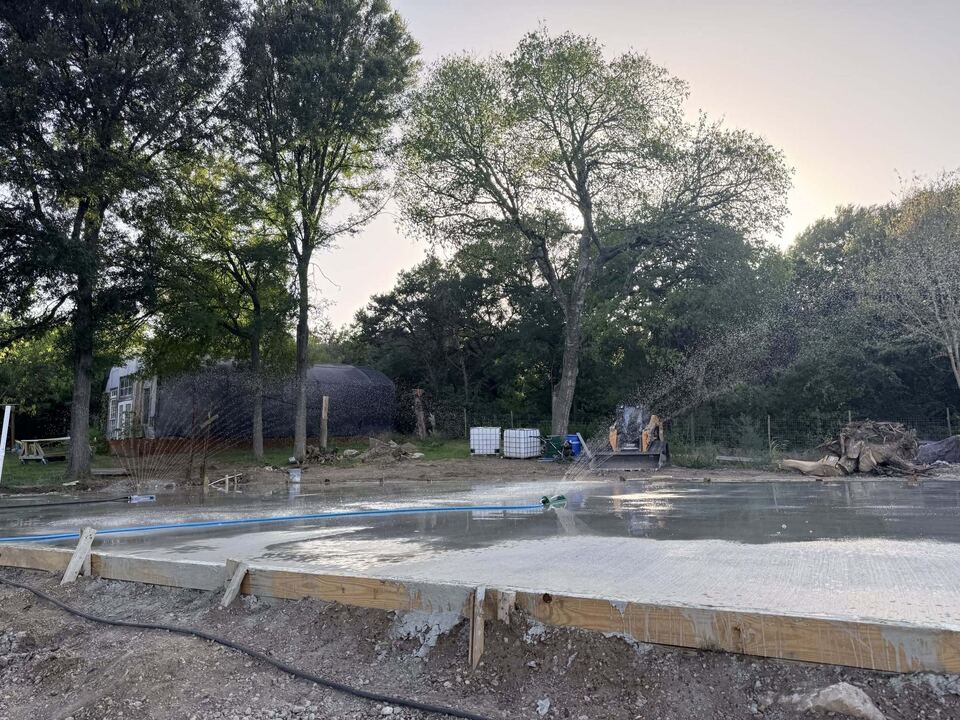 A freshly poured concrete foundation, its surface still wet
                  and reflecting the evening sky. Timber forms run along the
                  near edge, water lines cross the ground in front of it, and
                  a skid-steer sits parked at the far side beneath the
                  trees.