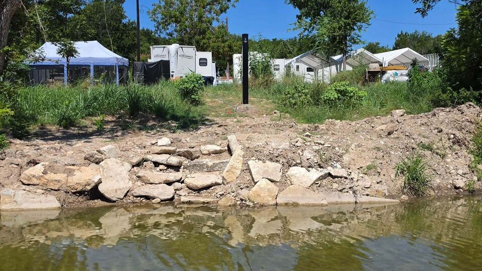The sensor tower seen from across the water, standing above a rebuilt stone bank, with travel trailers and a canopy among the trees behind.