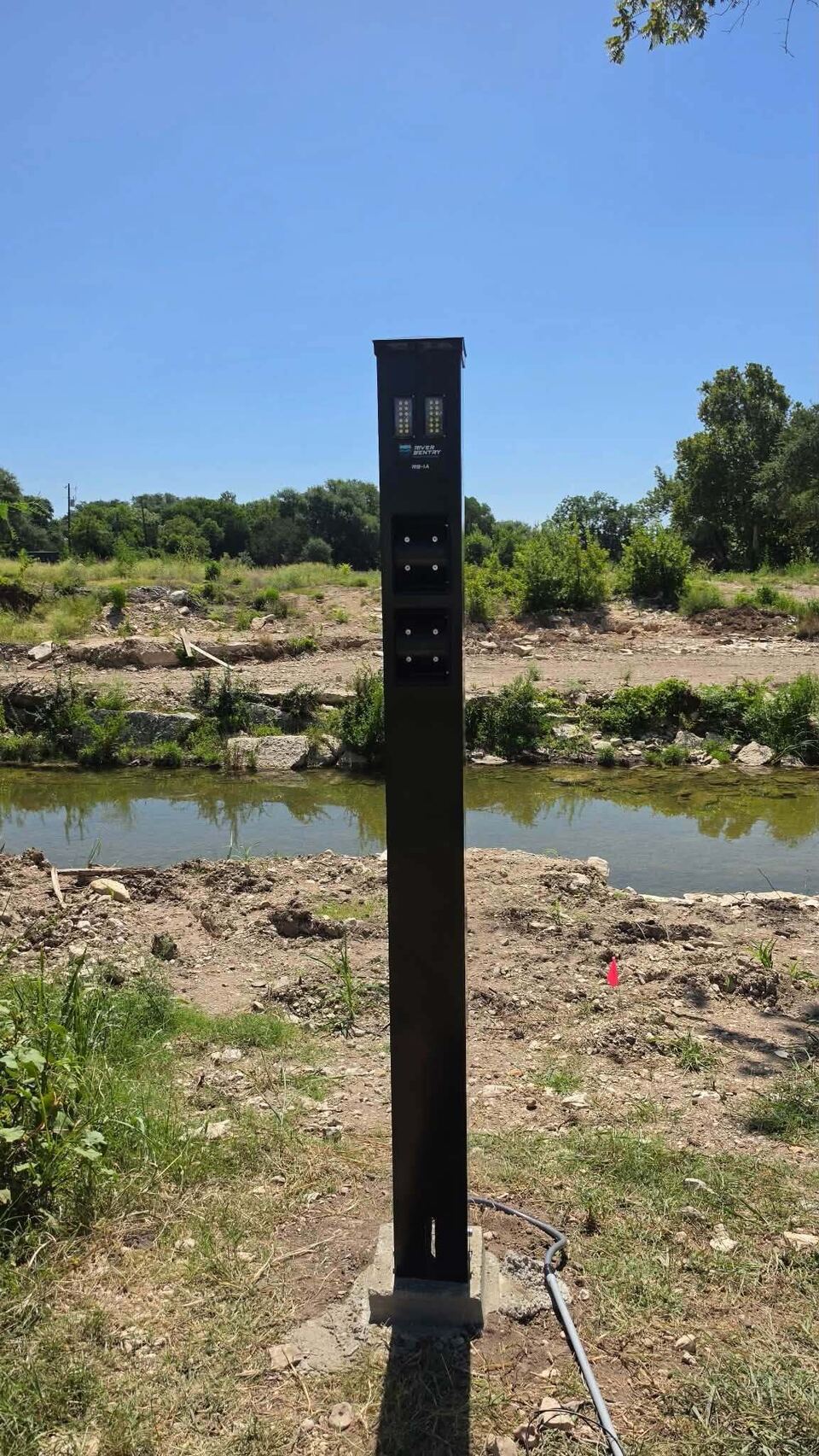 A tall black River Sentry sensor tower standing on the bank directly above Sandy Creek, with the scoured banks and clear water behind it under a blue sky.