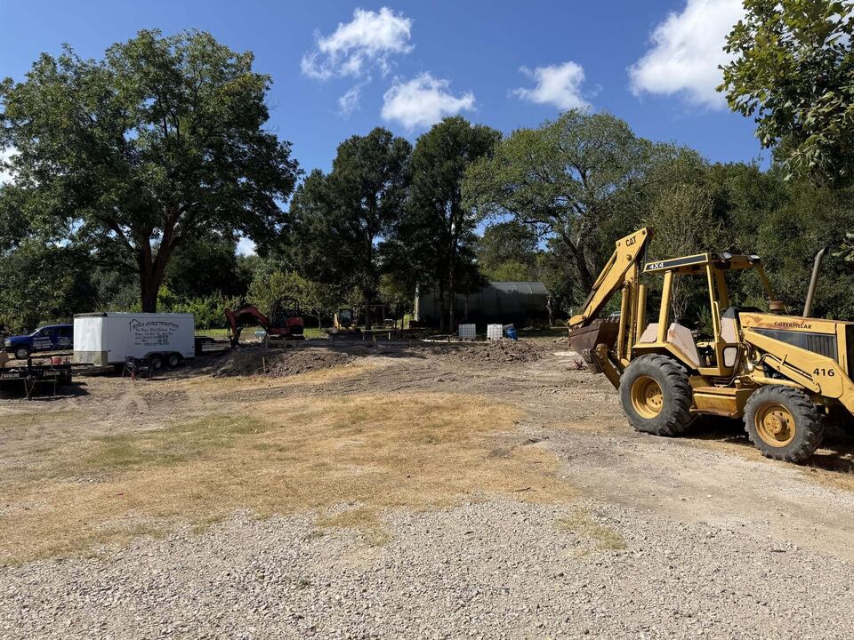 A yellow backhoe standing on newly graded gravel under a
                    blue sky, with a trailer and outbuildings along the
                    treeline behind it.